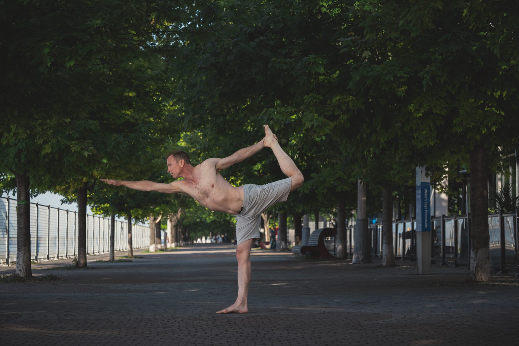 Christopher Gillett in dancer pose on a tree-lined waterfront promenade